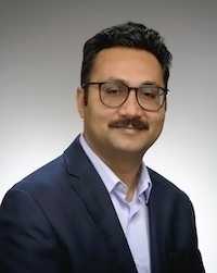 Headshot of a man with glasses, a mustache, and dark hair, wearing a dark blue blazer and light blue shirt against a gray background.