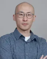 Bald East Asian man with glasses wearing a blue checkered shirt, looking forward with a neutral expression.